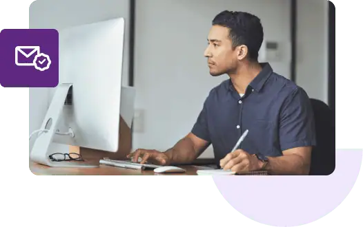 A man in a blue shirt types on his Mac with one hand and writes on a notepad with the other.