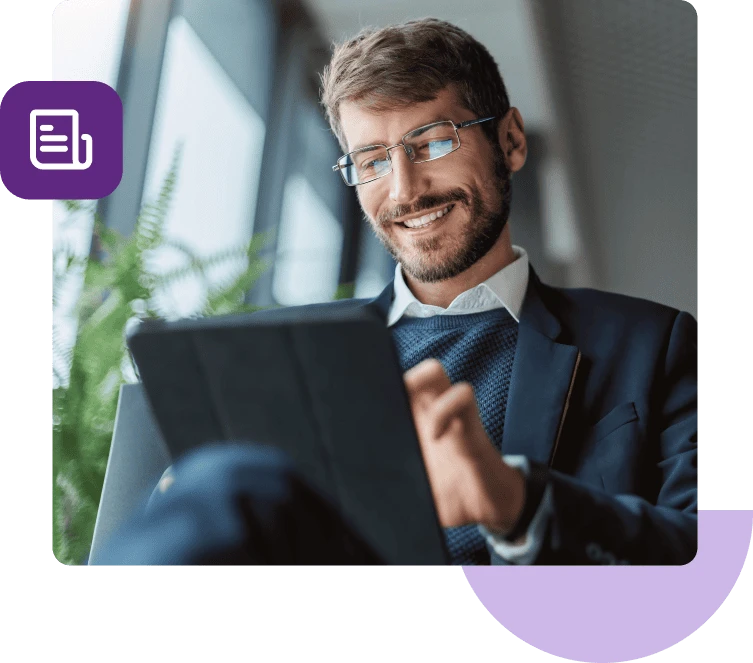 Un hombre con traje azul y gafas sentado frente a una planta sonríe mientras mira su tableta.