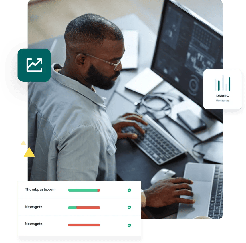 A man in a blue shirt works at his desk with one hand on his Mac keyboard and another on his laptop as he uses the DMARC checker to review compliance for a three domains.