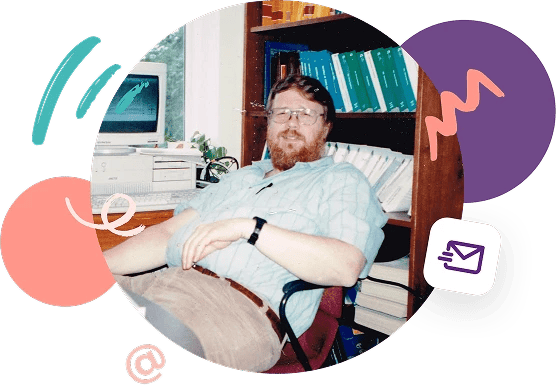 Computer programmer Ray Tomlinson, inventor of networked email, sitting at a desk and wearing glasses and a light blue short-sleeved shirt with bookcase in the background