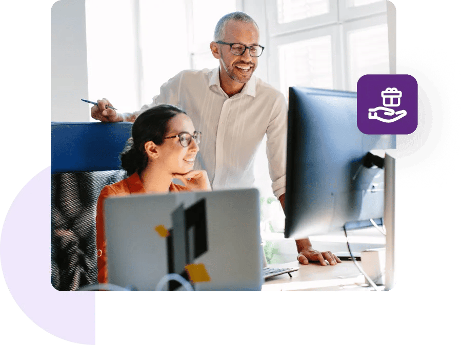 Man standing behind female coworker's chair, while both smile and look on at a desk monitor