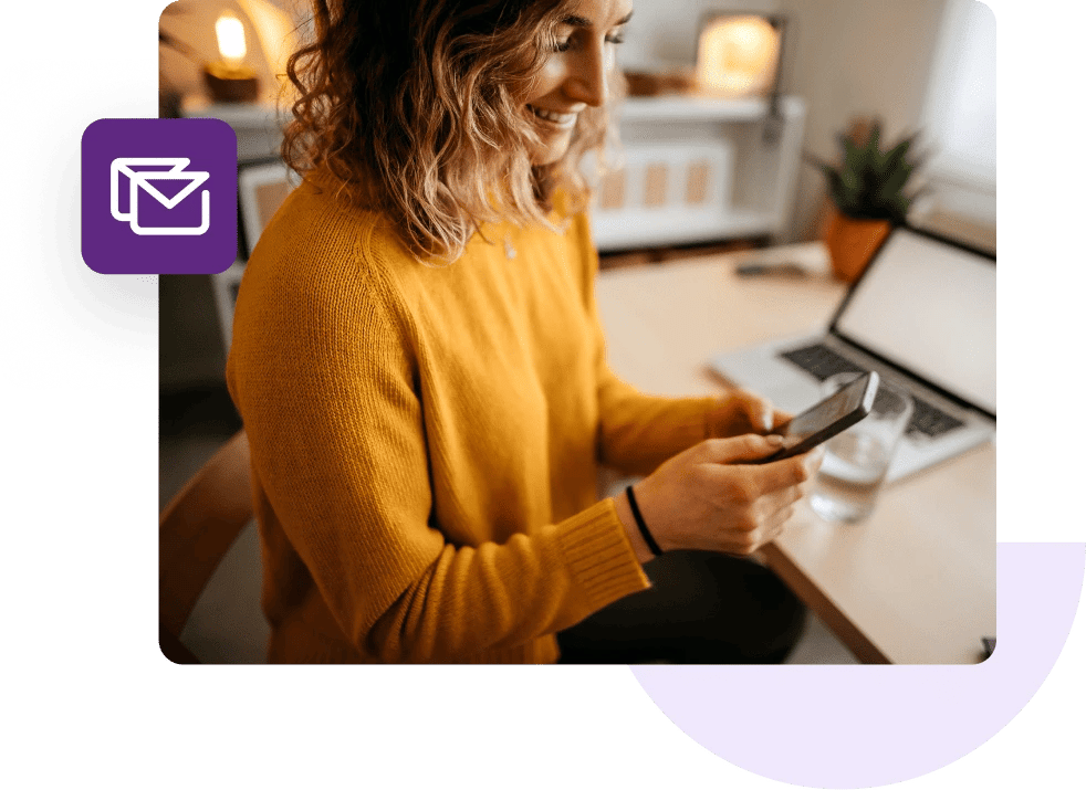 Woman sitting at her desk holding and looking at her phone with a laptop in the background