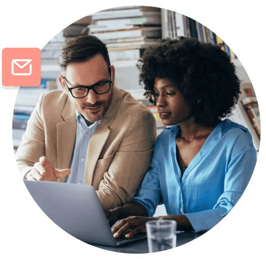 A man in a brown blazer and glasses sits shoulder to shoulder with a woman in a blue blouse as they discuss what they're looking at on a laptop.