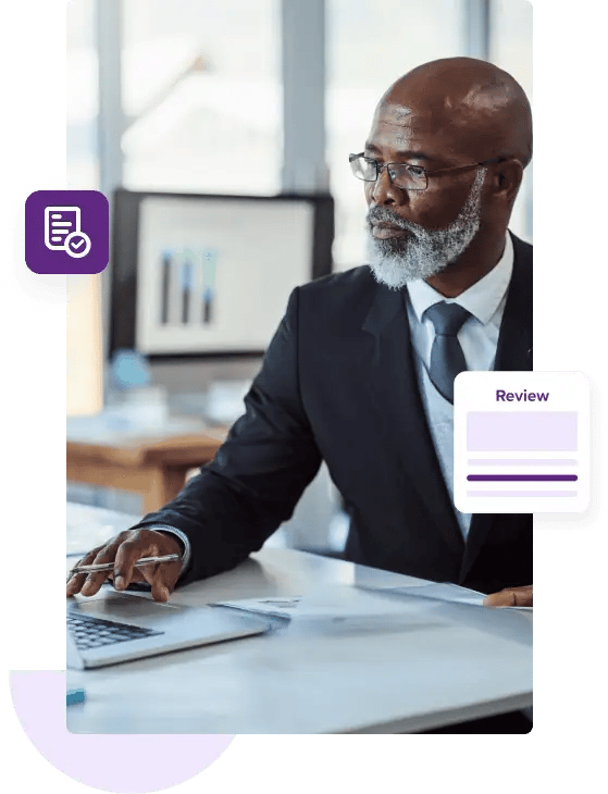 Un hombre de negocios calvo con barba gris se sienta en su escritorio vestido con un traje azul oscuro y extiende la mano para tocar la alfombrilla del mouse de su computadora portátil.