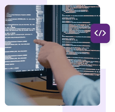 A developer sits in front of horizontal and vertical monitors with code and points at the horizontal one.