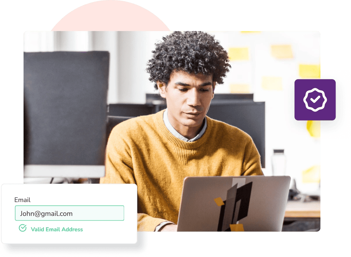 A man in an orange jumper sits in an office and types on his laptop to check if 'john@gmail.com' is a valid email address.