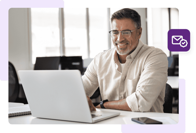 A man in glasses and a white shirt sits at a white desk and smiles in front of his open laptop.