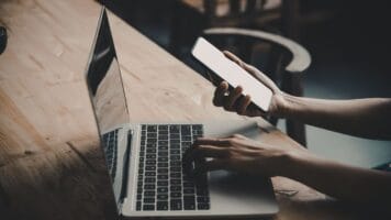 Marketer taps away at laptop while managing the smartphone to ensure great marketing.