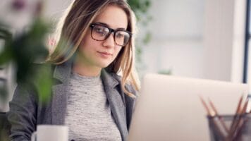 Young bespectacled woman sits in front of her laptop to set up email automation using her newly cleaned list.