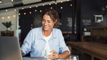 Hispanic woman clutches coffee cup and looks at laptop where she is performing an email health check.