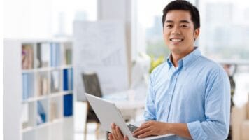 Young Asian man smiles while holding his laptop. He smiles because he knows his emails don't look like spam.