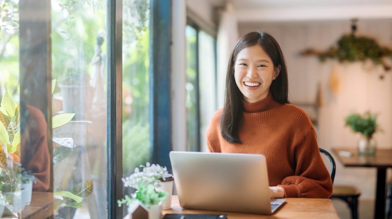Smiling Asian woman sits in front of laptop in well-lit room.