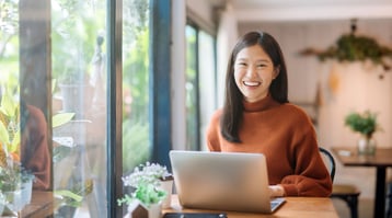 Smiling Asian woman sits in front of laptop in well-lit room.