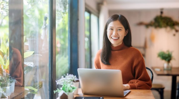 Smiling Asian woman sits in front of laptop in well-lit room.