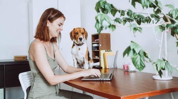 Solo woman entrepreneur wearing a light green dress smiling and working at laptop computer with small brown and white dog on her desk
