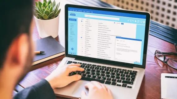 A person using a laptop with an open email inbox displayed on the screen, sitting at a desk with a notebook, glasses, and a potted plant.