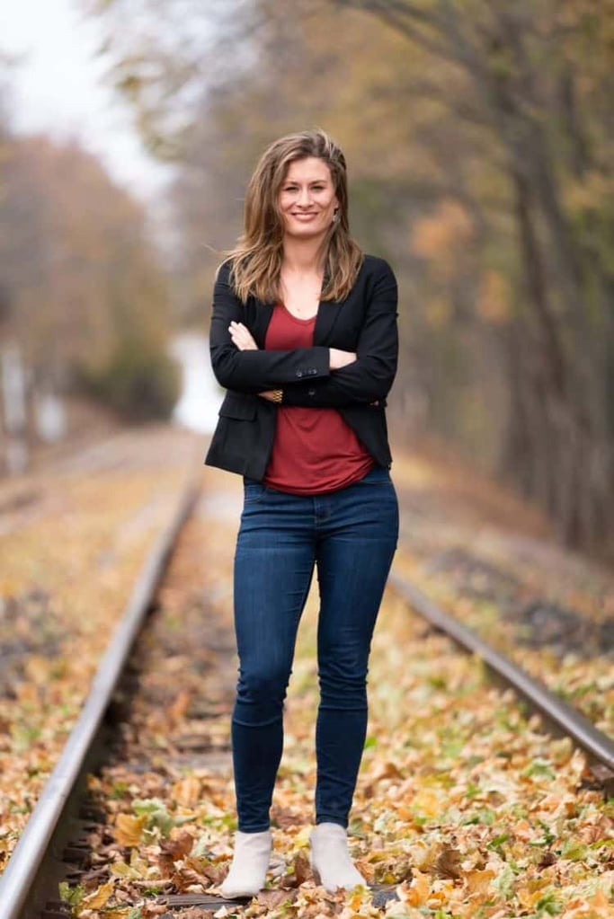 Image of Liz as she stands in the middle of train tracks with fall leaves all around her. She is smiling. 